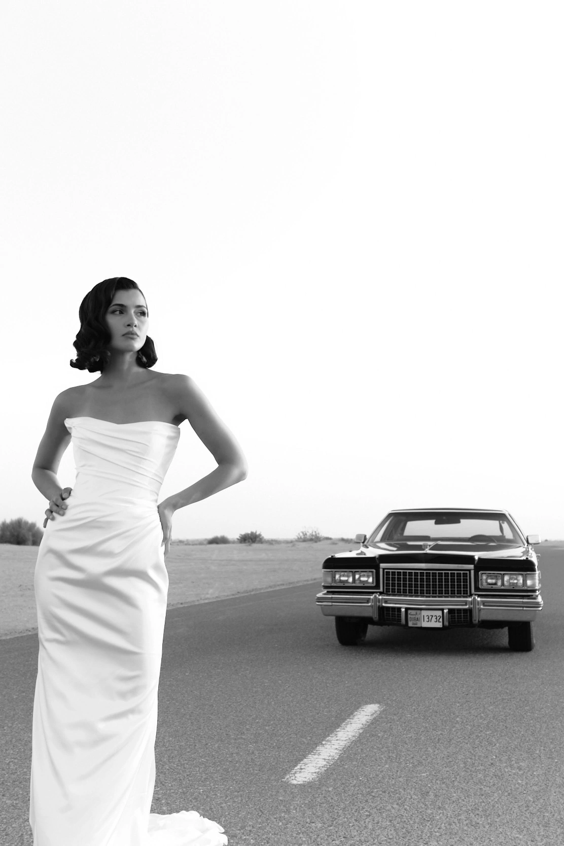 Desert Cadillac editorial, Dubai: bride in a white gown on an open desert road, a vintage black Cadillac behind her, black and white