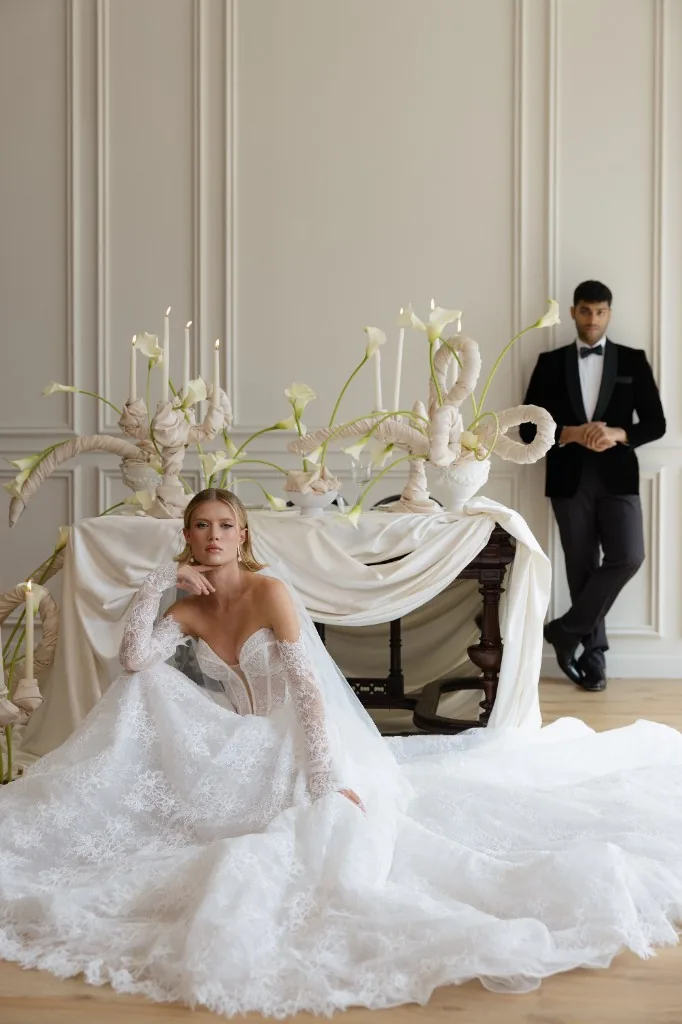 Editorial wedding portrait in Dubai: bride in lace gown seated on wood floor, groom in black velvet, cream table with calla lilies and taper candles